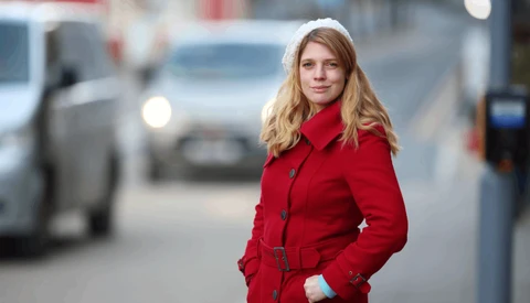 Woman in red coat standing on urban street with blurred cars in background.