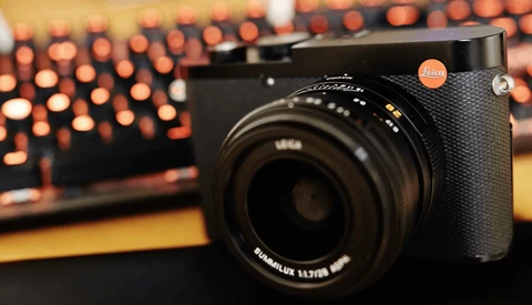 Leica Q2 camera with black leather grip and Summilux lens on wooden surface with blurred bokeh lights in background.