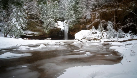 Frozen river with cascading waterfall framed by snow-covered evergreen trees and rocky cliffs.