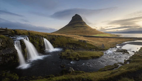 Twin waterfalls cascade into a dark river with a distinctive cone-shaped mountain rising behind moss-covered terrain.