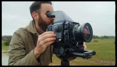 Cinematographer operating a film camera with viewfinder and attached lens in an open field.