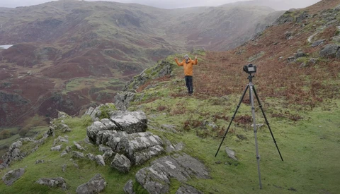 Surveyor with theodolite on tripod in mountainous valley landscape.