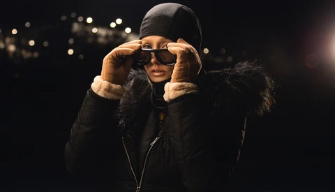 Person wearing dark winter jacket and sunglasses adjusting glasses against dark background with warm bokeh lights.