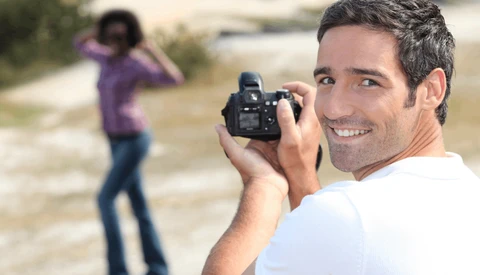 Male photographer smiling at camera while holding a DSLR, with female subject posing in background on rural path.
