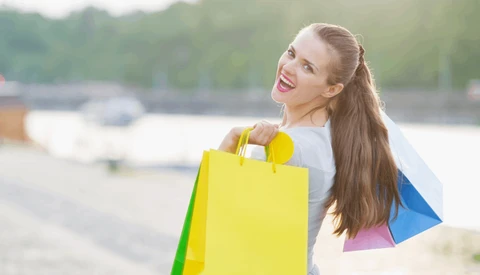 Woman holding colorful shopping bags at a waterfront location, smiling at camera.