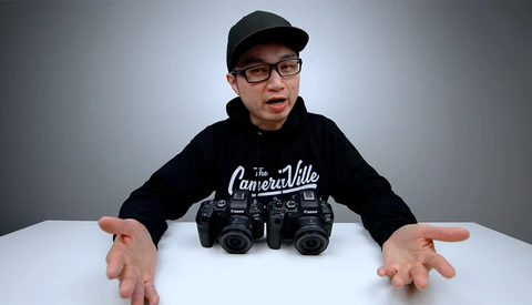 Young photographer wearing glasses and black cap displays two professional camera bodies on white table.