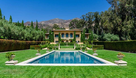 Symmetrical formal garden with rectangular swimming pool, manicured lawn, and yellow villa framed by cypress trees and mountains.