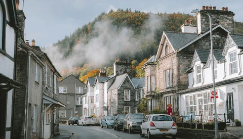 Misty mountains loom behind a quiet village street lined with parked cars and traditional stone buildings.