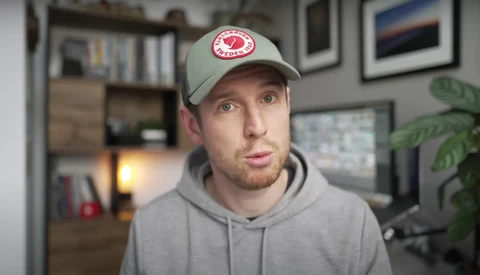 Man wearing gray hoodie and light cap with red circular logo, speaking directly to camera in home office setting.