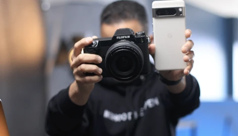 Person holding a black Fujifilm camera in one hand and a white smartphone in the other.