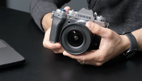 Hands holding a Fujifilm mirrorless camera with attached lens over a dark surface.