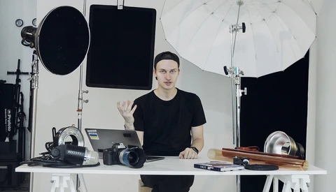 Photographer gesturing while seated at desk surrounded by professional camera equipment and lighting gear.