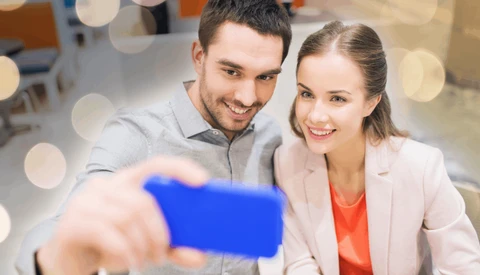 Two professionals smiling while taking a selfie with a smartphone in an office setting.