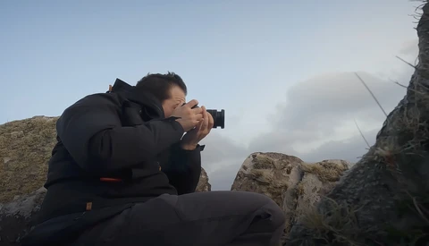 Photographer crouched on rocky terrain, holding a camera to their face while composing a shot against a cloudy sky.