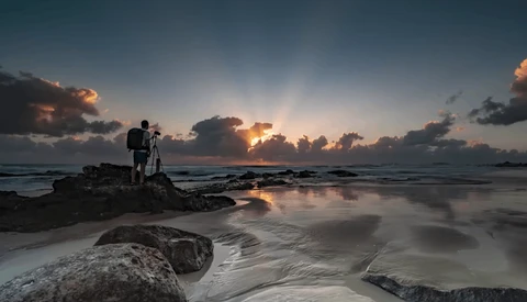 Photographer with tripod silhouetted on rocky outcrop during golden hour coastal sunset.