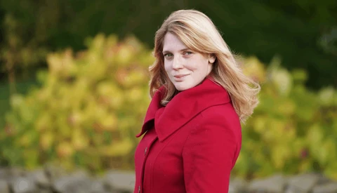 Woman with blonde hair wearing a red blazer, photographed outdoors against blurred green foliage.