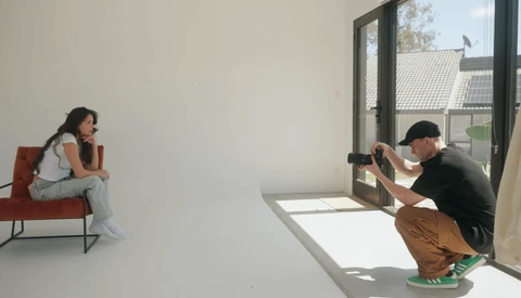 Photographer crouching while shooting a model seated on a red chair in a minimalist white studio.