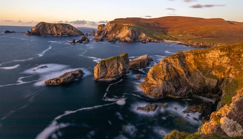 Rocky sea stacks illuminated by golden hour light along a rugged coastline with rolling hills.