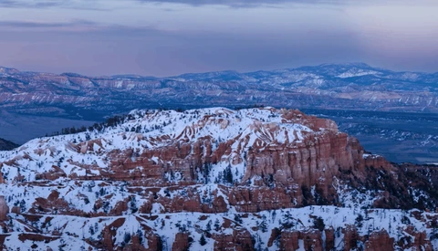 Snow-covered hoodoo formations and ridges in red rock canyon landscape with distant mountains under twilight sky.