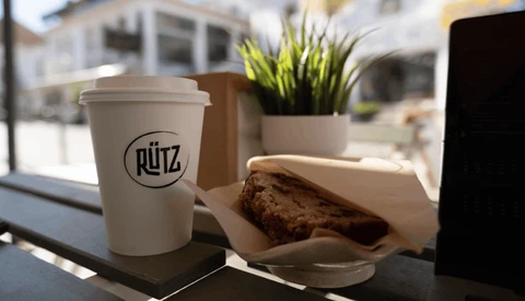 Coffee cup with Rütz logo beside pastry on windowsill with potted plant and blurred street view.