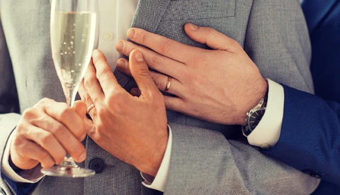 Two people wearing wedding rings hold champagne glasses while touching hands.