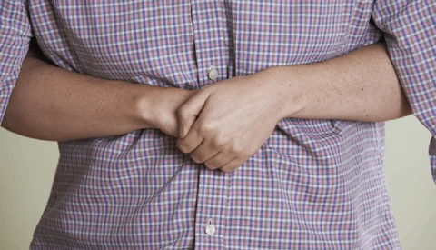 Person in checkered shirt with hands clasped across stomach in studio setting.