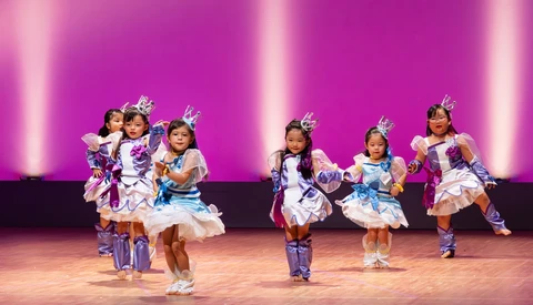 Young dancers in purple and white costumes perform on a stage with pink backlighting.