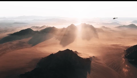 Aerial view of a desert landscape with mountains silhouetted against golden sunlight and dust clouds.