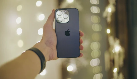 Hand holding a dark blue iPhone with quad camera setup against blurred warm-toned bokeh background.