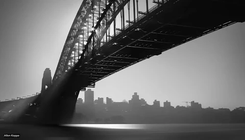 Steel arch bridge silhouetted against a misty cityscape reflected in still water below.