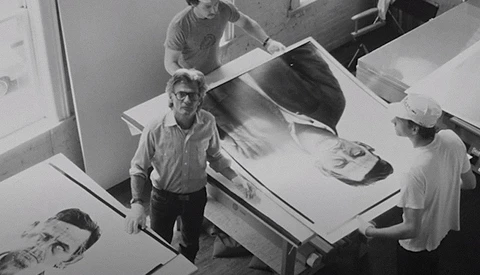 Black and white overhead view of four people examining large photographic prints on a work table.