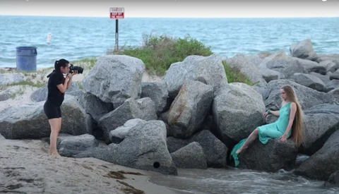 Photographer shooting a mermaid-tailed model posed on large boulders at a beach.