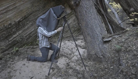 Photographer kneeling behind tripod-mounted camera in rocky cave setting, with dark cloth covering head and camera.