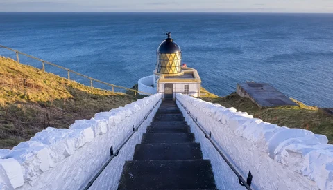 A symmetrical staircase with white railings leads to a yellow lighthouse with a black domed top on a coastal cliff.