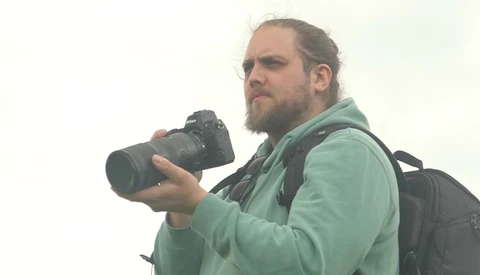 Male photographer holding a DSLR camera with telephoto lens, wearing a green jacket and backpack.