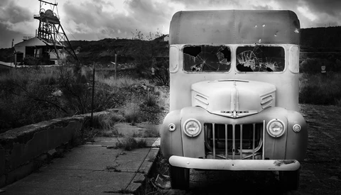 Abandoned truck with broken windows at a derelict mining site with headframe visible in background.