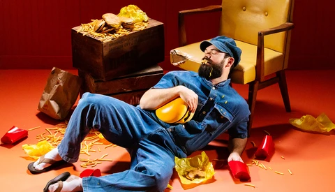 Man in denim lying on orange floor surrounded by gold coins and coins spilling from boxes against red background.