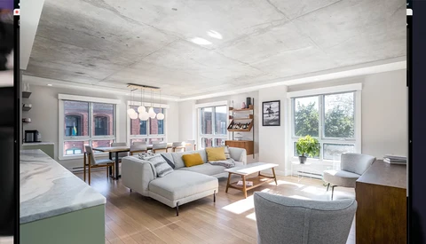 Bright modern open-concept loft with light wood floors, concrete ceiling, and abundant natural light from large windows.