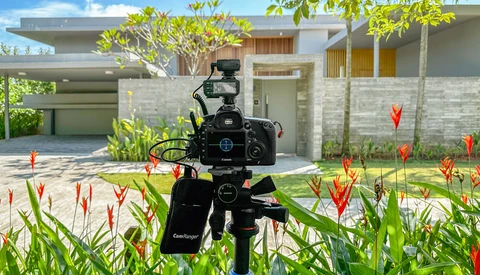 Professional video camera on tripod filming a modern residential house surrounded by tropical plants.
