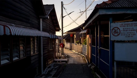 Narrow alley lined with dark wooden buildings at dusk, with warm golden light illuminating the street and distant figures walking.