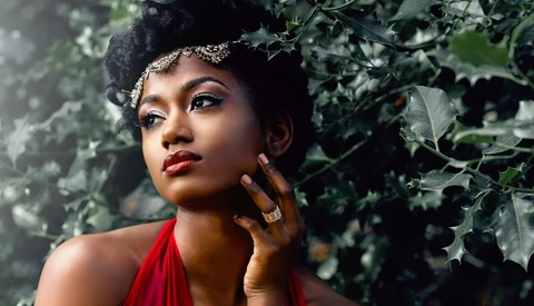 Woman with bold red lips and winged eyeliner posed against ivy-covered wall, touching her face thoughtfully.