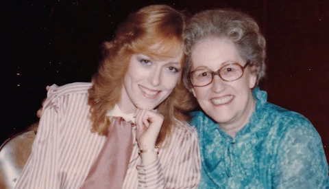 Two women smiling at the camera in a close portrait composition against a dark background.