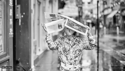 Person wearing patterned shirt holds newspapers above their head on a city street.