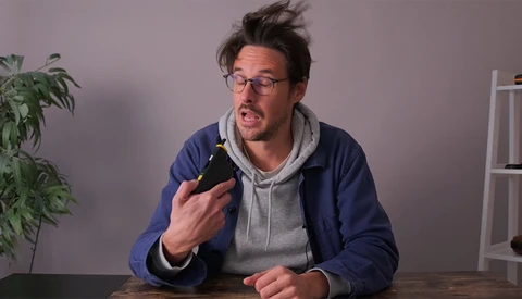 Man in glasses holding a dark bottle while speaking animatedly at a table.