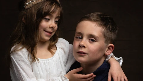 Young girl in white dress with tiara and boy in dark shirt posed together against dark background.