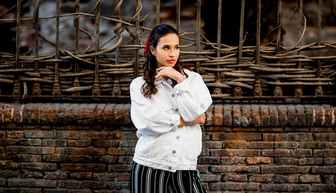 Woman in white button-up shirt and striped skirt posing against weathered brick wall with woven branches.