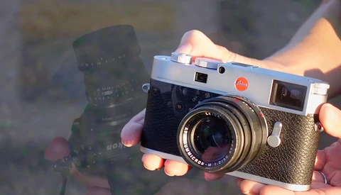 Hands holding a silver rangefinder camera with a dark telephoto lens in the background, backlit by warm golden light.