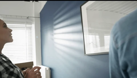 Two people in an interior space with blue walls and bright window light creating linear shadows across the surface.