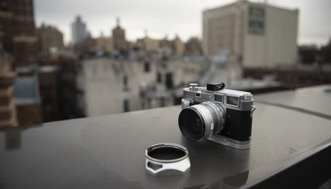 Vintage rangefinder camera with lens cap displayed on a rooftop overlooking an urban cityscape.