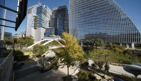 Modern urban plaza with curved white architectural feature, high-rise office buildings, and mature trees under clear blue sky.